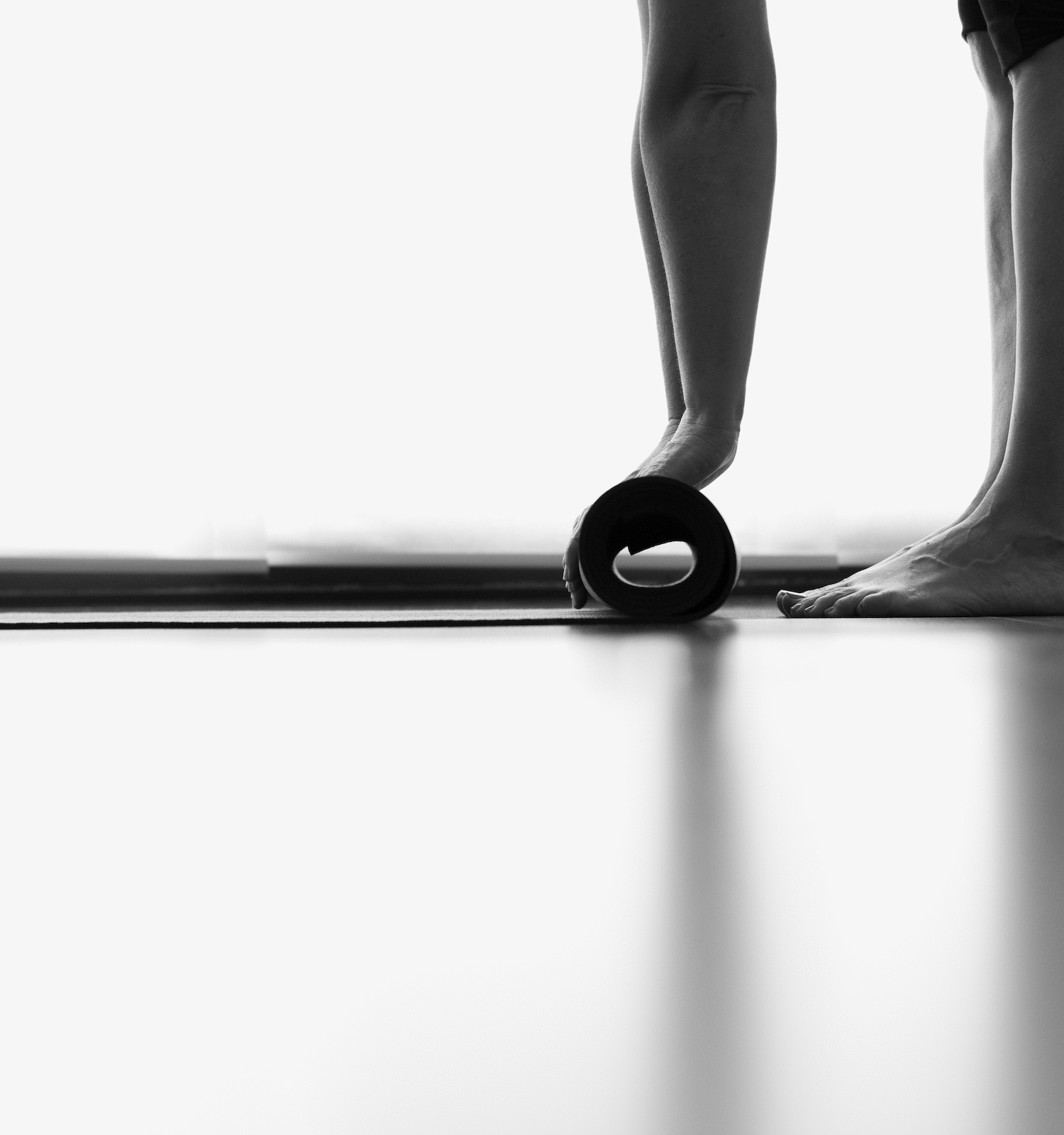 A person is seen rolling up a yoga mat on the floor, with only their legs and feet visible in a minimalistic, low-light setting.