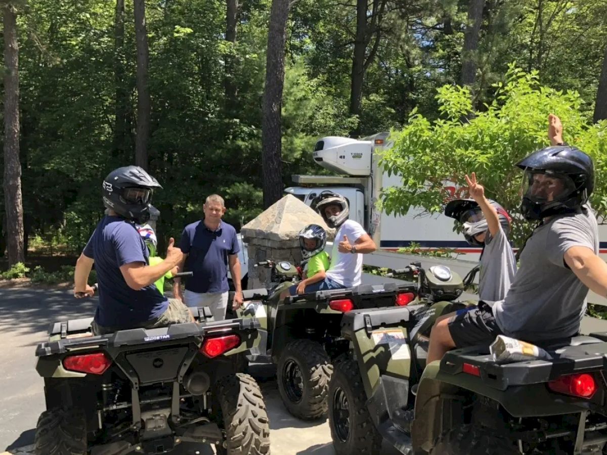 A group of people, some wearing helmets, are gathered around ATVs in a wooded area with a person standing nearby.