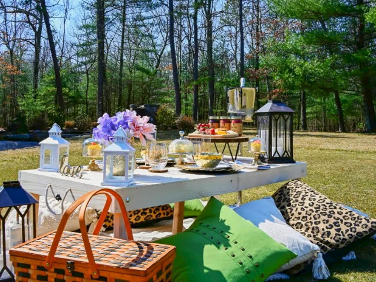 A picnic setup on a low table with various foods, lanterns, flowers, pillows, and a wicker basket, set in a wooded area.