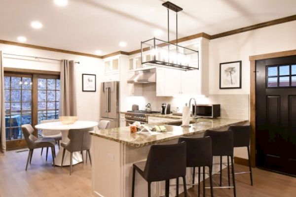 A modern kitchen with a center island, stools, a dining table, stainless steel appliances, white cabinets, framed artwork, and a black front door.