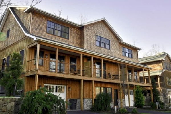 The image shows a large, multi-story house with a wooden exterior, multiple balconies, and garages on the lower level. It has a rustic design.