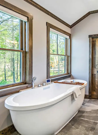 A modern bathroom with a freestanding bathtub, double-sink vanity, large mirror, and large windows with wooden trim and stone tile flooring.