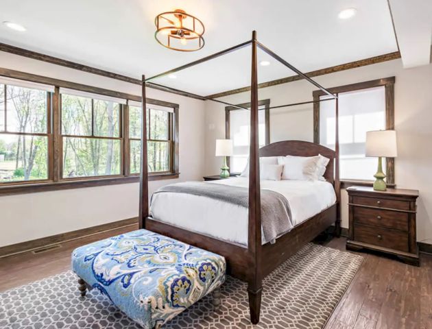 A bedroom with a canopy bed, wooden furniture, large windows, a patterned ottoman, a rug, and two lamps on bedside tables, ending the sentence.