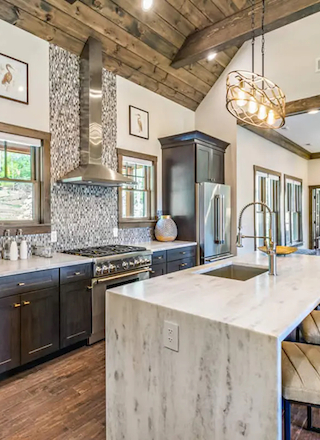 A modern kitchen with dark cabinets, a white island, stainless-steel appliances, bar stools, pendant lights, and a stone accent wall ending the sentence.