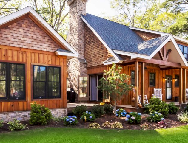 The image shows a charming wooden house with a stone chimney, a front porch with rocking chairs, and a beautifully landscaped garden.