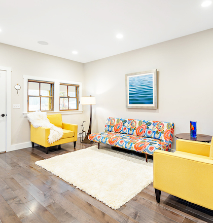 A brightly lit living room with yellow chairs, a colorful sofa, a white rug, wall art, and wood flooring, featuring windows and a door.