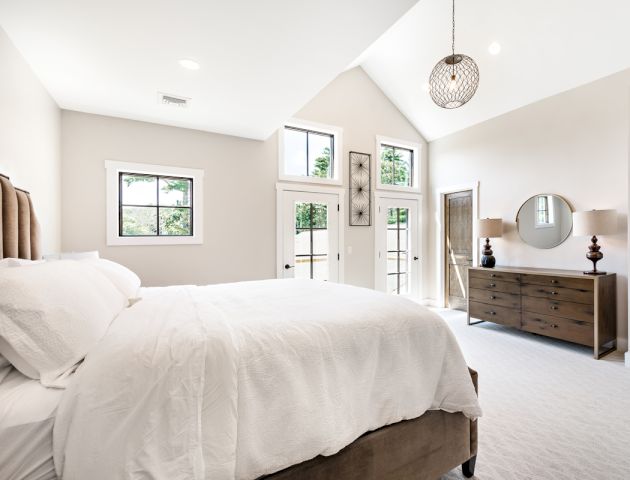 A bright, modern bedroom with a large window, white bed, wooden dresser with lamps and a round mirror, and white walls.