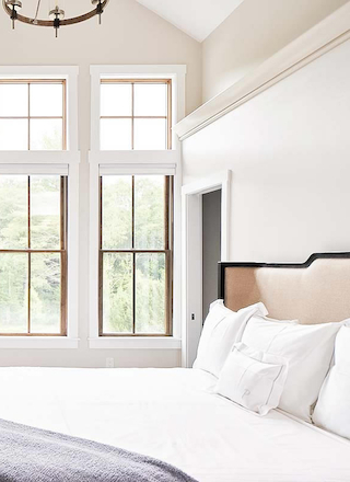 A bright bedroom with tall windows, white walls, a modern chandelier, built-in storage, a bed with white bedding, and a gray blanket at the foot.