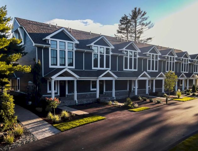 A row of modern two-story townhouses with a neat front yard and garden, situated along a paved driveway, and surrounded by trees.