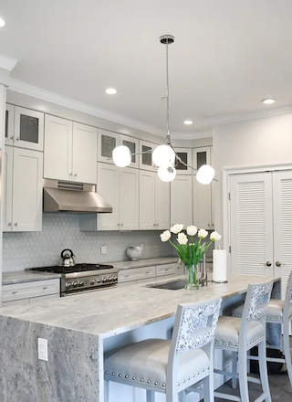 A modern kitchen with white cabinets, a stainless steel refrigerator, a marble island with chairs, pendant lights, and white flowers on the counter at Hilltop Lodge