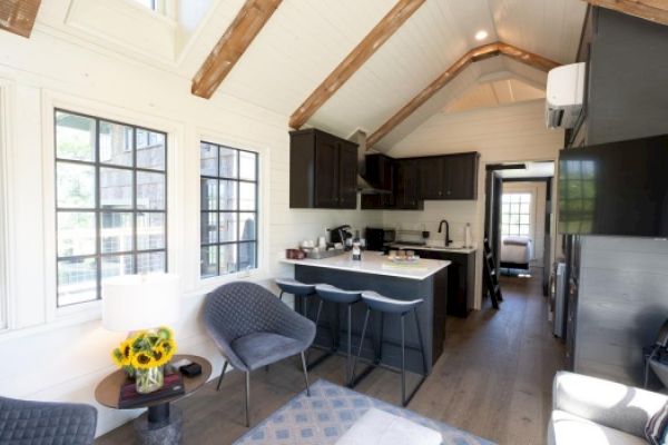 The image shows a cozy, modern tiny home interior with a kitchen, sitting area, large windows, and exposed ceiling beams.