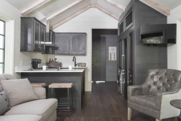 A modern, compact living space with a small kitchen, dark cabinets, seating area with sofa and armchair, and vaulted ceiling with exposed beams.