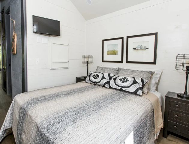 A cozy bedroom with a bed, gray bedding, pillows, two side tables with lamps, wall-mounted TV, and framed pictures above the bed.