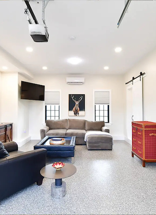 A bright, modern room features a gray couch, a black armchair, a TV, wall art, sliding barn doors, and air conditioning, all on a polished floor.