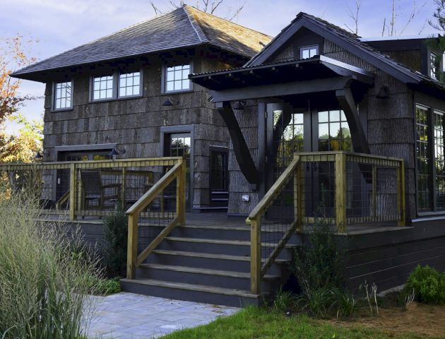 A modern, wooden house with large windows, a wrap-around porch, and lush greenery in the yard, situated in a serene environment.