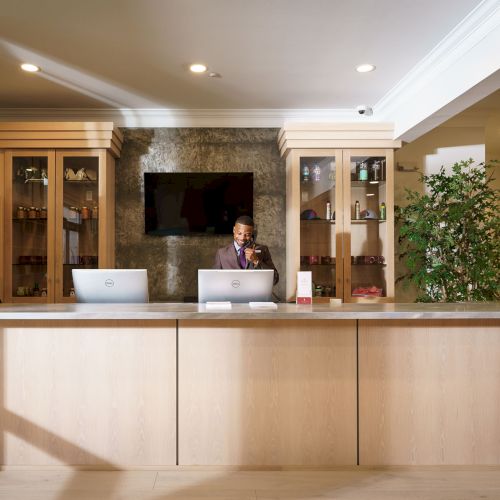 This image shows a reception desk with two computers, a receptionist handling a phone call, and decor including plants and artwork on the wall.