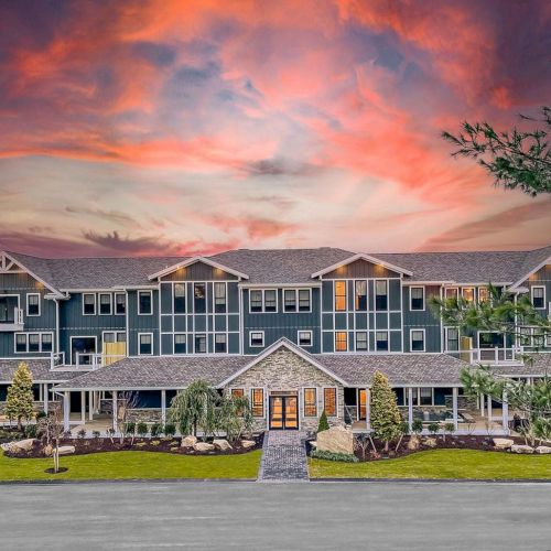 A large, modern building with multiple windows is set against a dramatic sunset backdrop, featuring landscaped grounds and a central pathway ending the sentence.