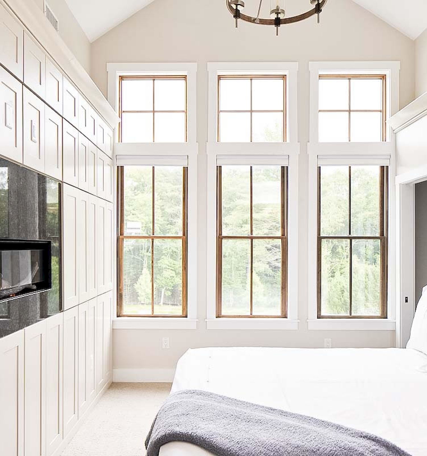 A bright bedroom features a large bed, built-in cabinetry with a TV, and three large windows letting in natural light.