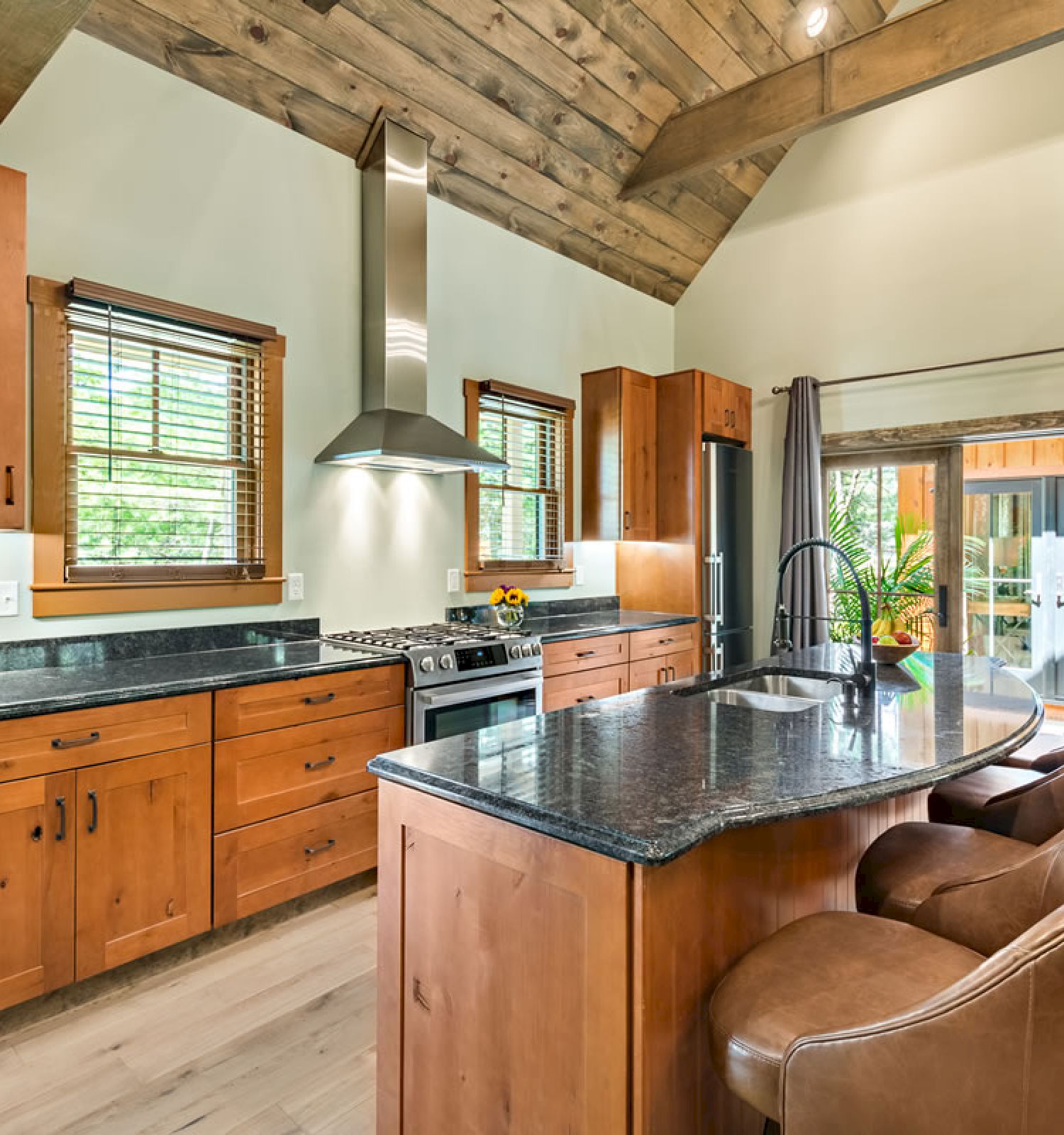 The image shows a modern kitchen with wooden cabinets, a large island, stainless steel appliances, and an open view to a patio area through sliding doors.