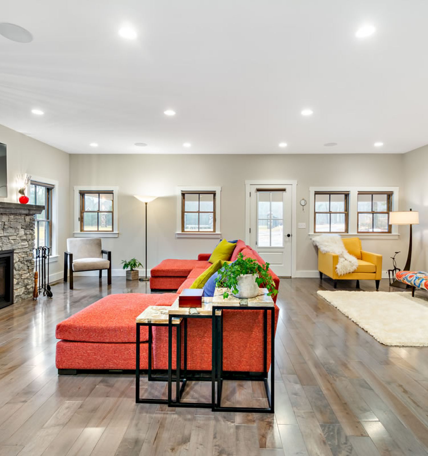 A spacious living room with a fireplace, colorful furniture, hardwood floors, and a wall-mounted TV, well-illuminated by ceiling lights.