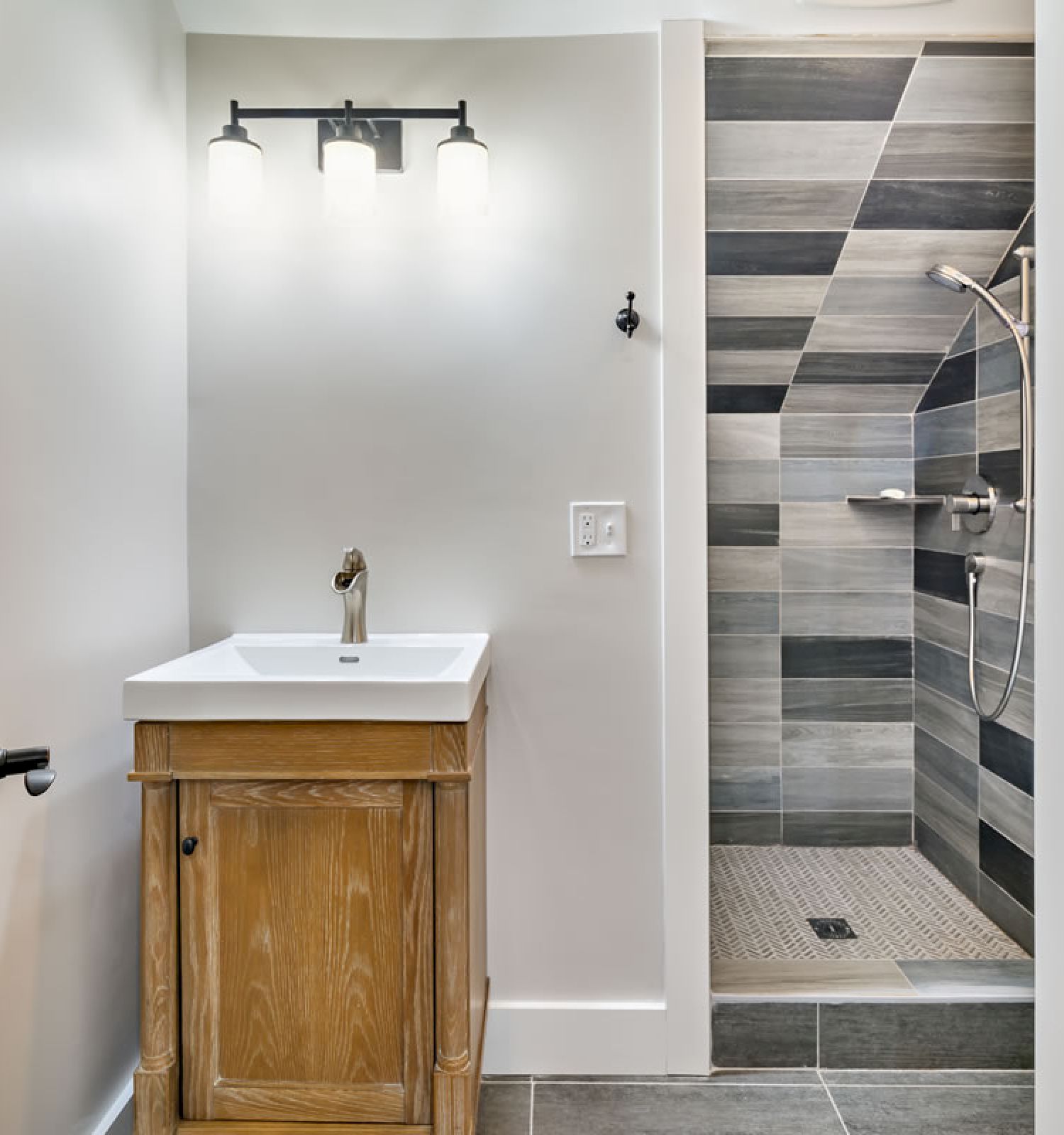 A well-lit bathroom with a wooden door, a wooden vanity with a sink, and a modern tiled shower with a glass door on the right wall.