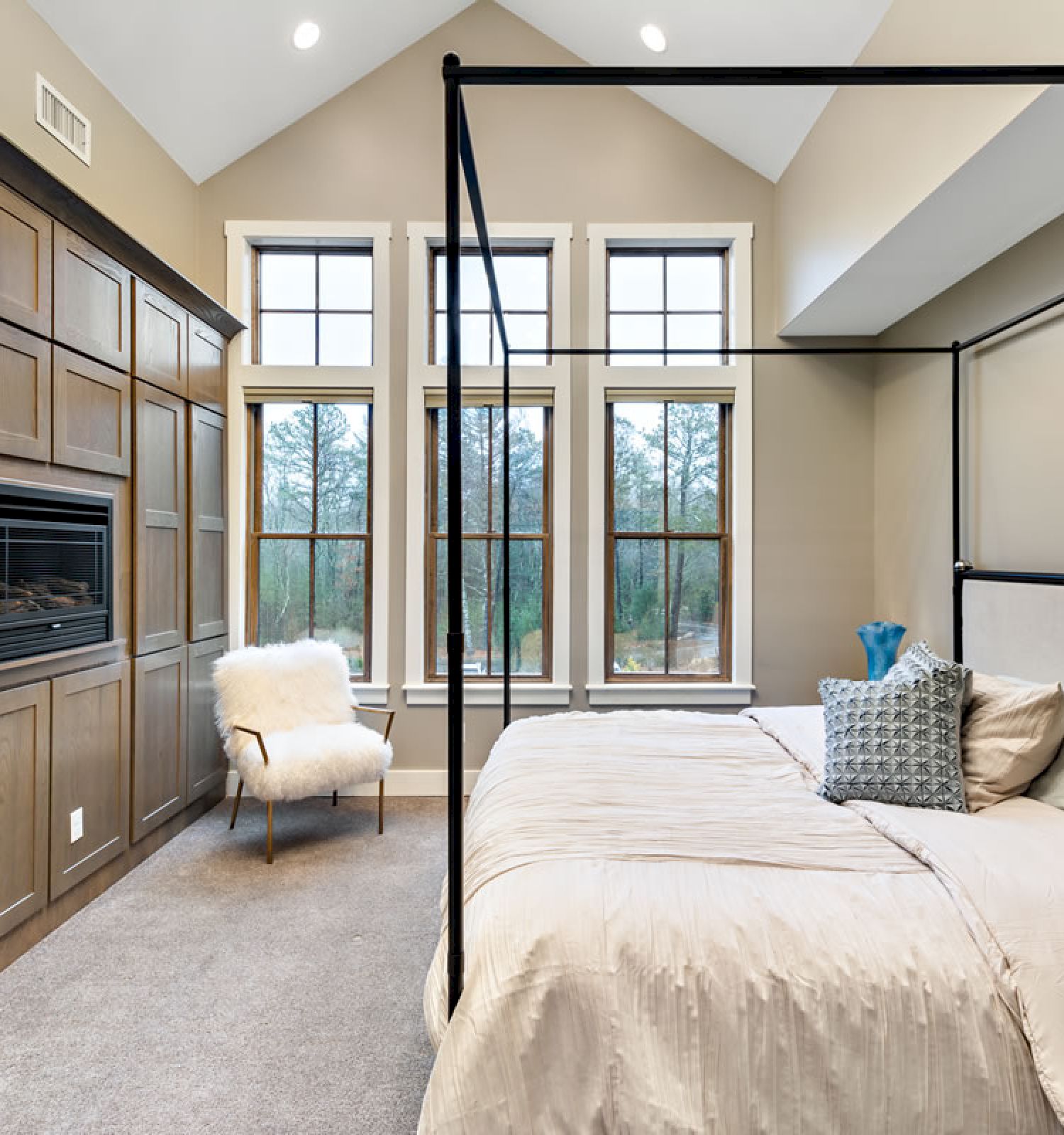 A modern bedroom with a canopy bed, large windows, built-in wooden cabinets, a fireplace, and a white chair near the windows, neutral décor.