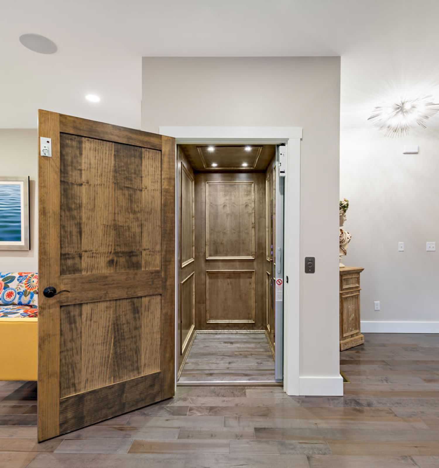 A modern interior features a wooden elevator with an open door, a colorful couch with playful pillows, a plush rug, and hardwood floors.