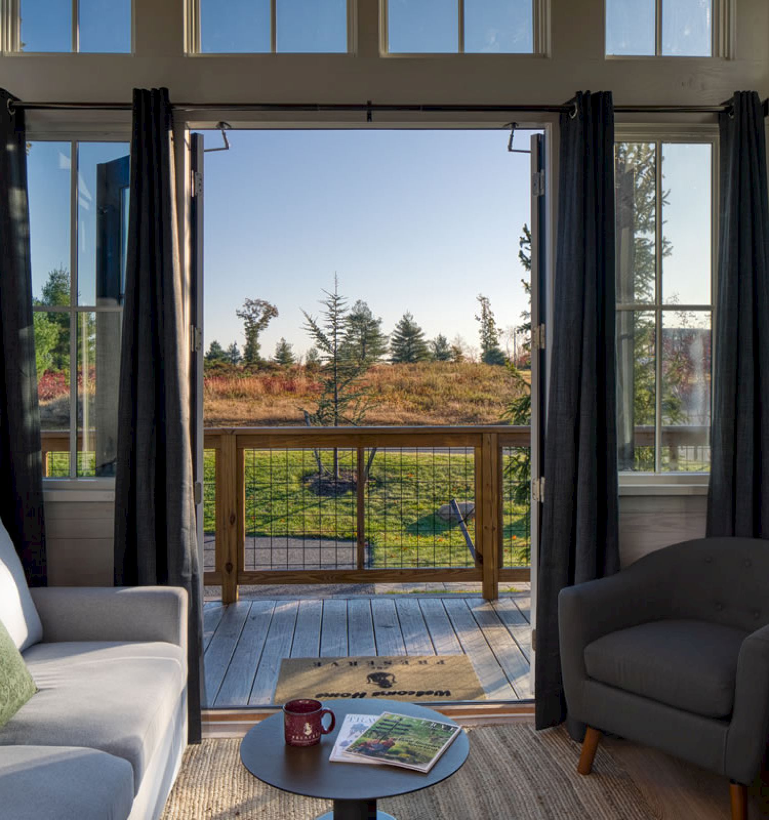 A cozy living room with a gray sofa, armchair, lamp, coffee table, and open doors leading to a wooden deck overlooking a scenic outdoor view.