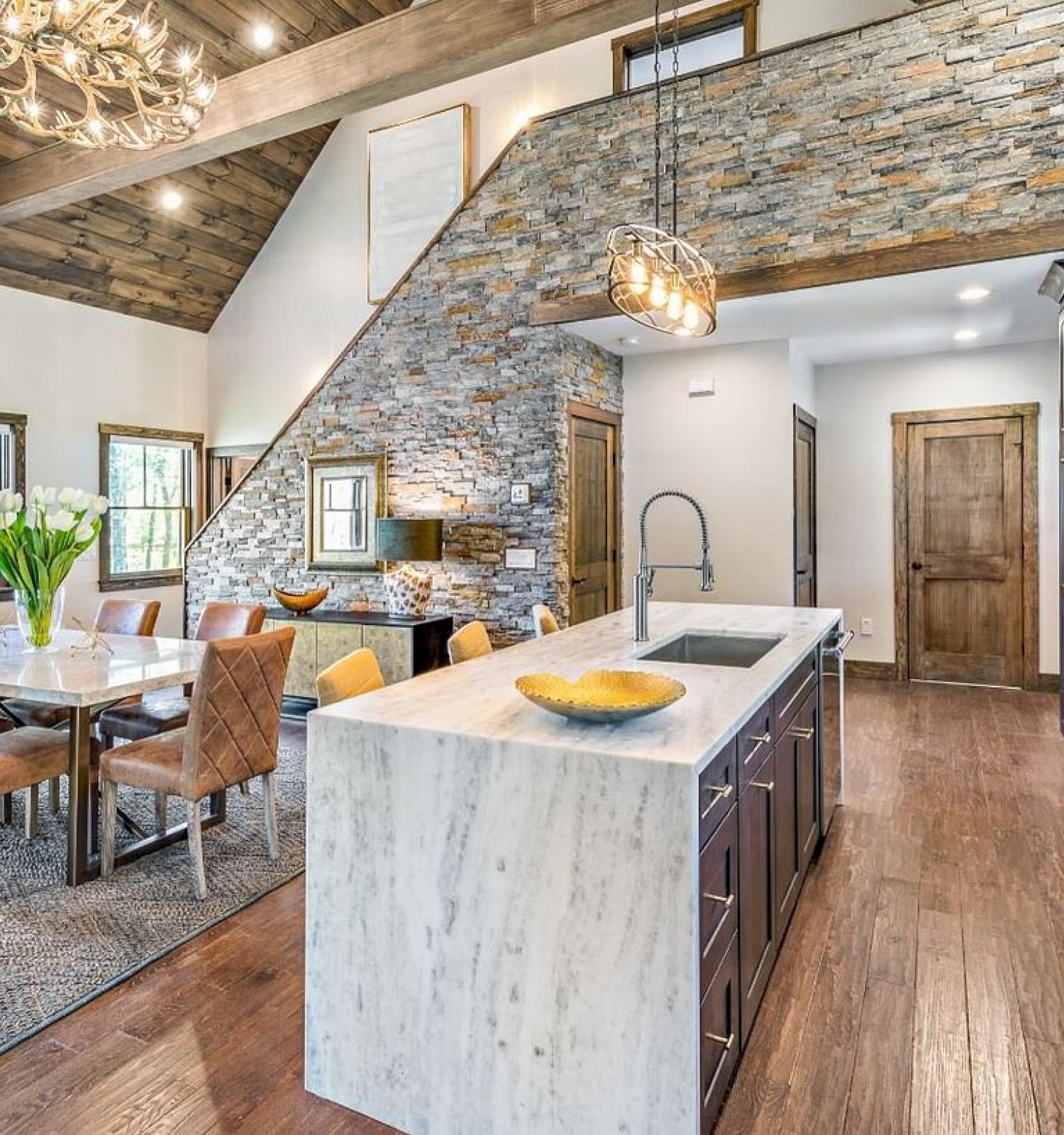 This image shows a modern kitchen and dining area with a marble island, leather chairs, wooden accents, and a high, beamed ceiling with hanging lights.