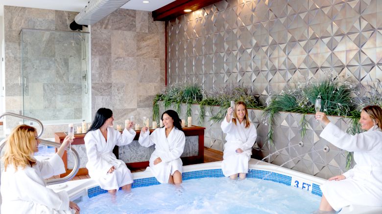 Five people in white robes are sitting around a hot tub, raising glasses in a toast. The space has modern decor and wall plants.