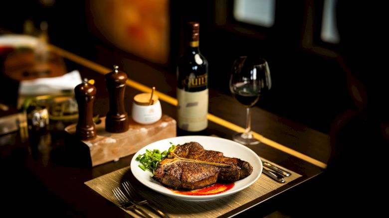 A plate with steak and greens is on a table next to a bottle of wine, a glass, and seasoning containers on a wooden surface.