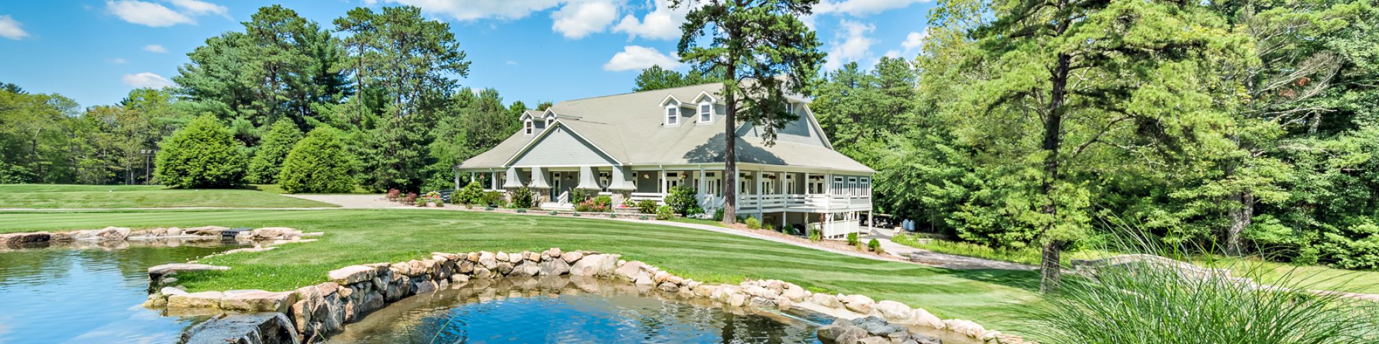 A serene landscape features a pond, lush greenery, tall trees, and a large white house in the background under a bright blue sky with scattered clouds.