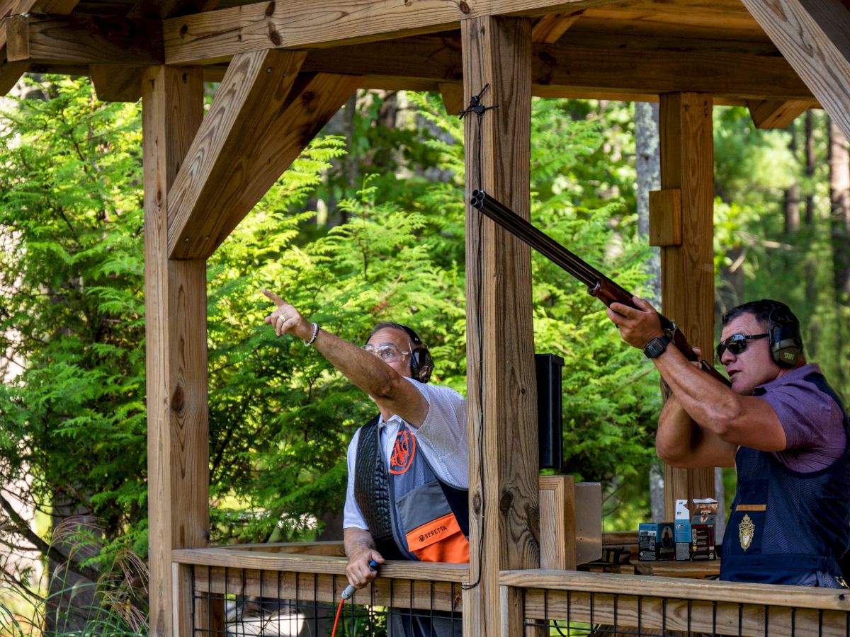 Two people are at an outdoor shooting range with headphones on; one is aiming a shotgun while the other points and gives instructions.