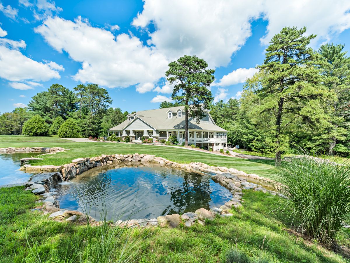 A large house surrounded by lush greenery and trees, with a small pond in the foreground under a blue sky with scattered clouds ending the sentence.