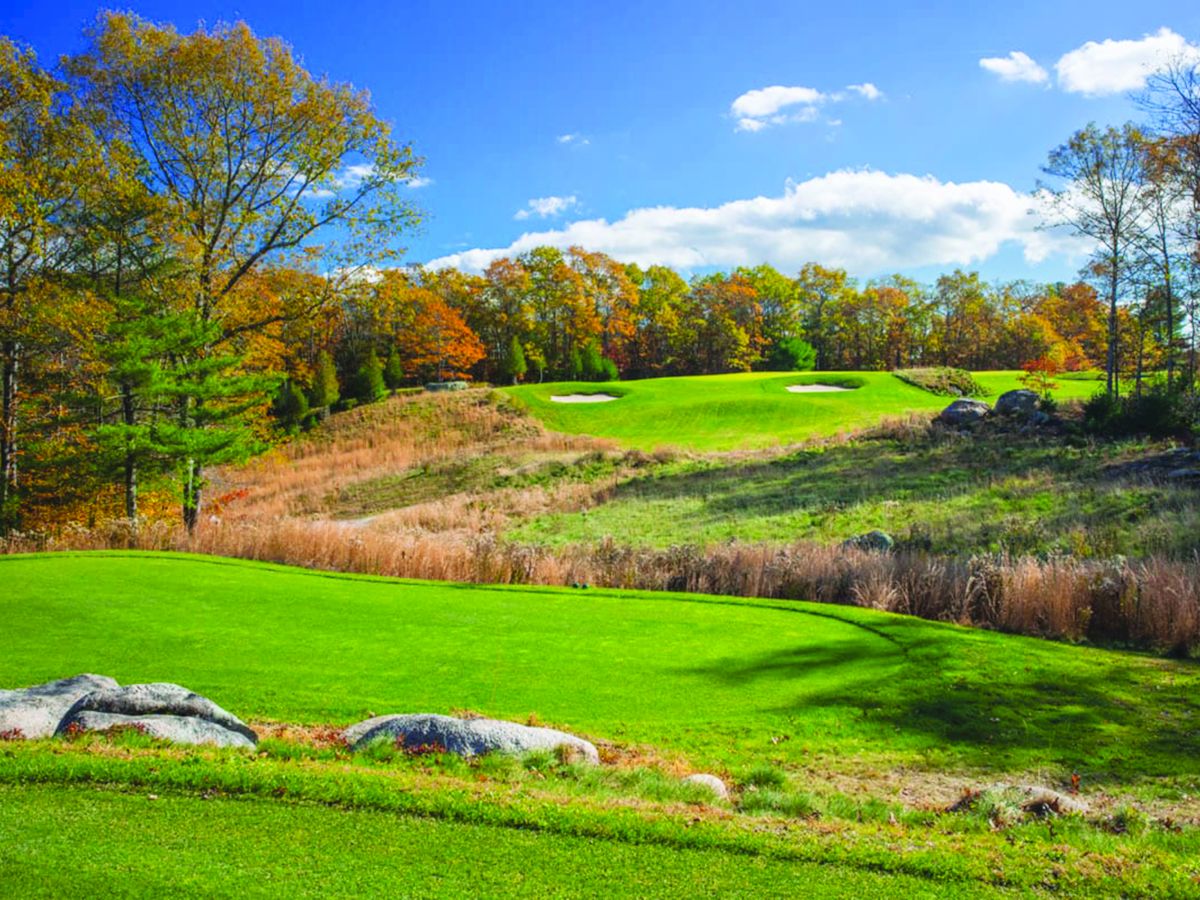 The image shows a golf course with green fairways, sand bunkers, trees in autumn colors, and a clear blue sky with scattered clouds.