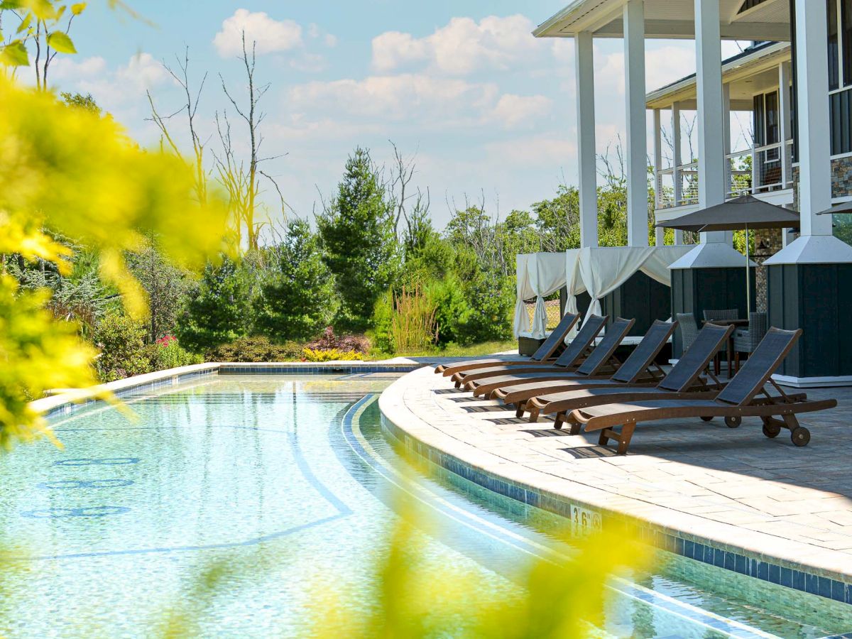 The image shows a serene poolside scene with several lounge chairs, cabanas, and lush greenery under a partly cloudy sky.