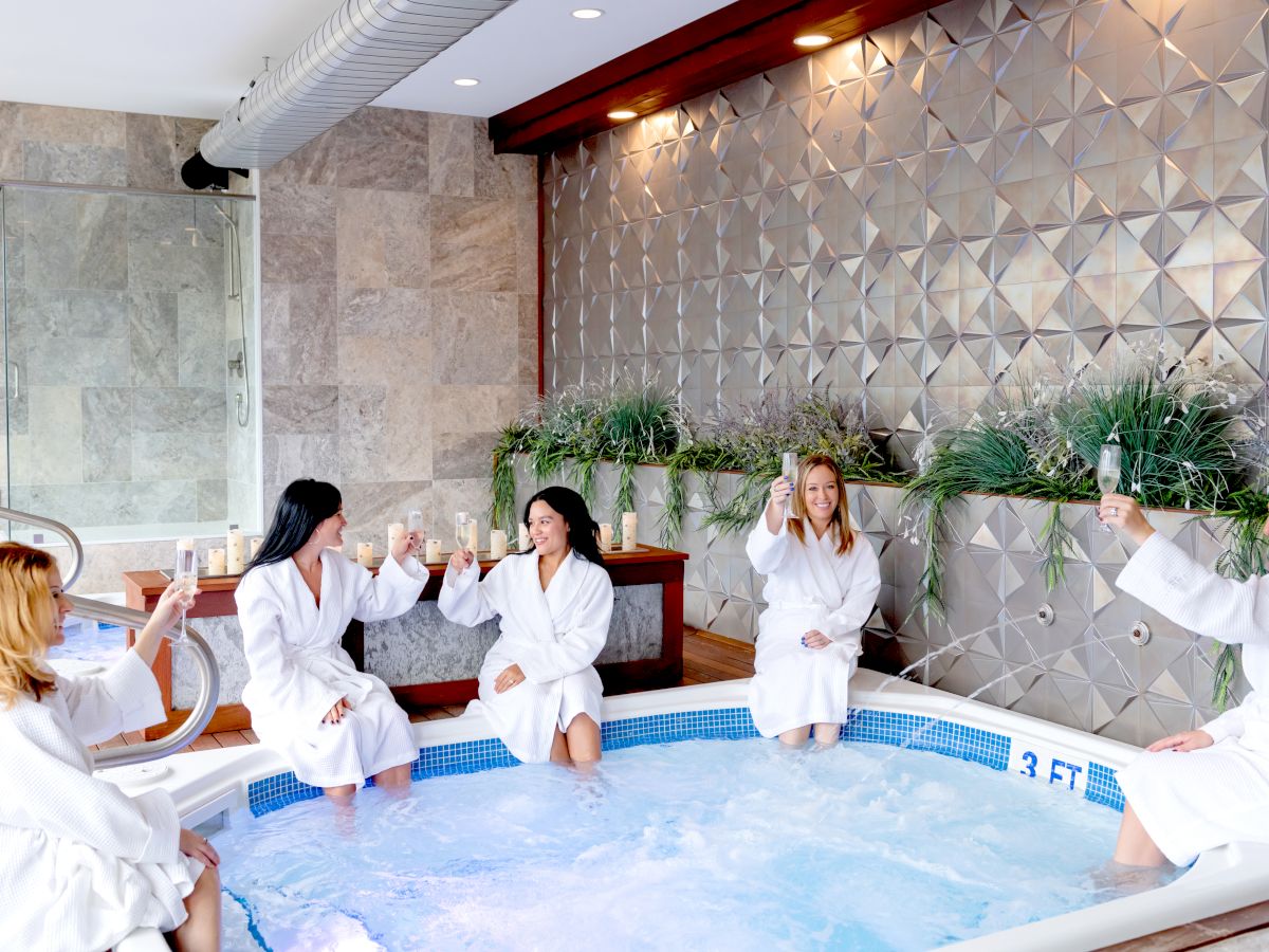 Five women in white robes are sitting on the edge of a hot tub, raising their glasses in a toast, in a spa-like setting.