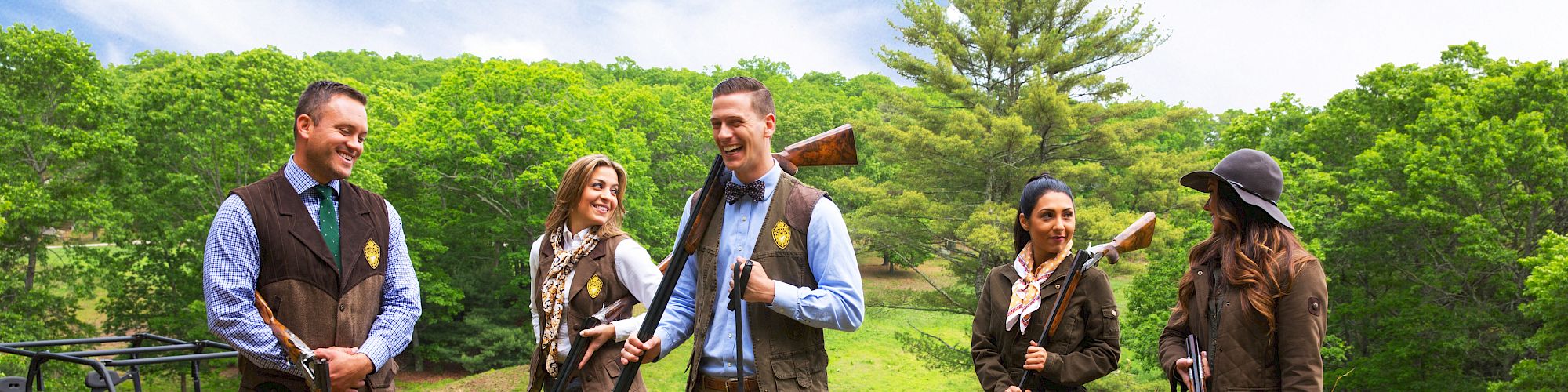 A group of five people and a dog are outside with hunting gear, wearing vests and casual outdoor clothing, with wooded scenery in the background.