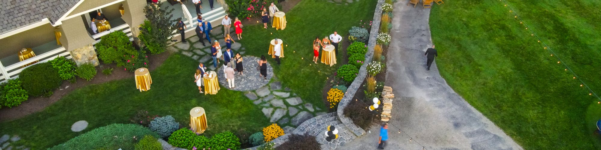 An aerial view of a gathering with people outside a large building with green lawns and pathways, surrounded by trees and landscape design.