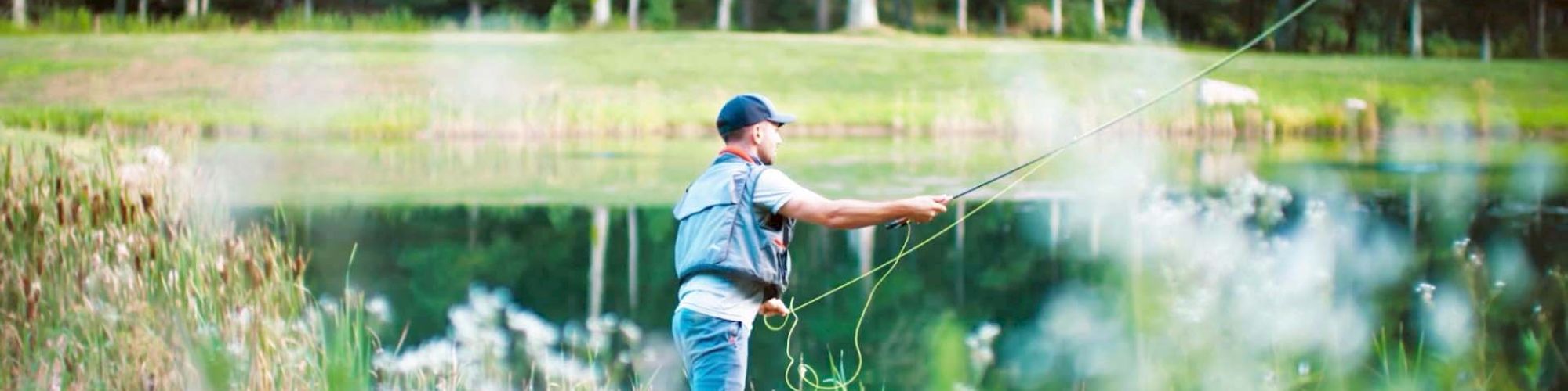 A person is fly fishing by a lake surrounded by greenery and trees, with a fishing rod in hand and casting a line into the water.