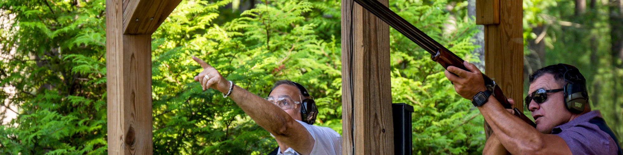 Two people at a shooting range, one pointing while the other aims a shotgun. Both are wearing ear protection, surrounded by greenery under a wooden structure.