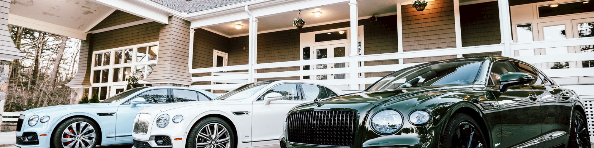 The image shows three luxury cars parked in front of a two-story house with an expansive porch and multiple windows, all bathed in daylight.