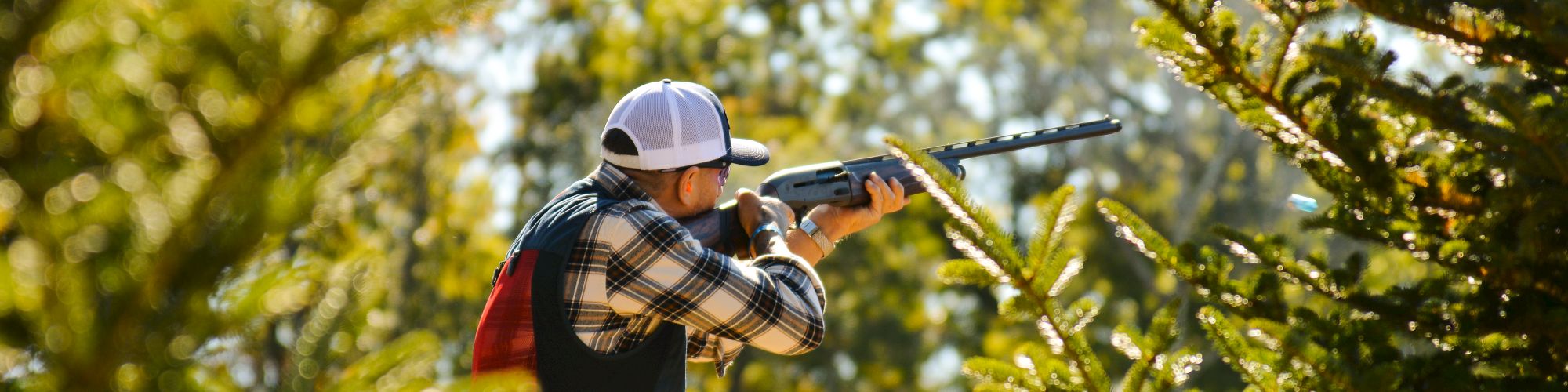 A person wearing a plaid shirt and cap aims a rifle from a wooden platform surrounded by trees outdoors.