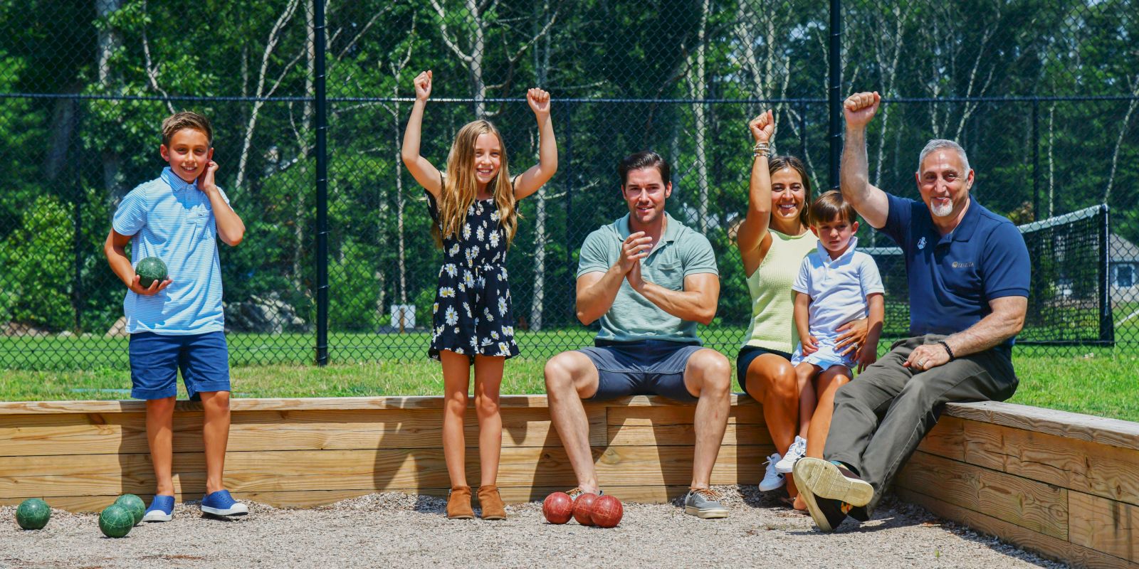 A group of five people—three children and two adults—celebrate while playing bocce ball in an outdoor court surrounded by trees and a fence.