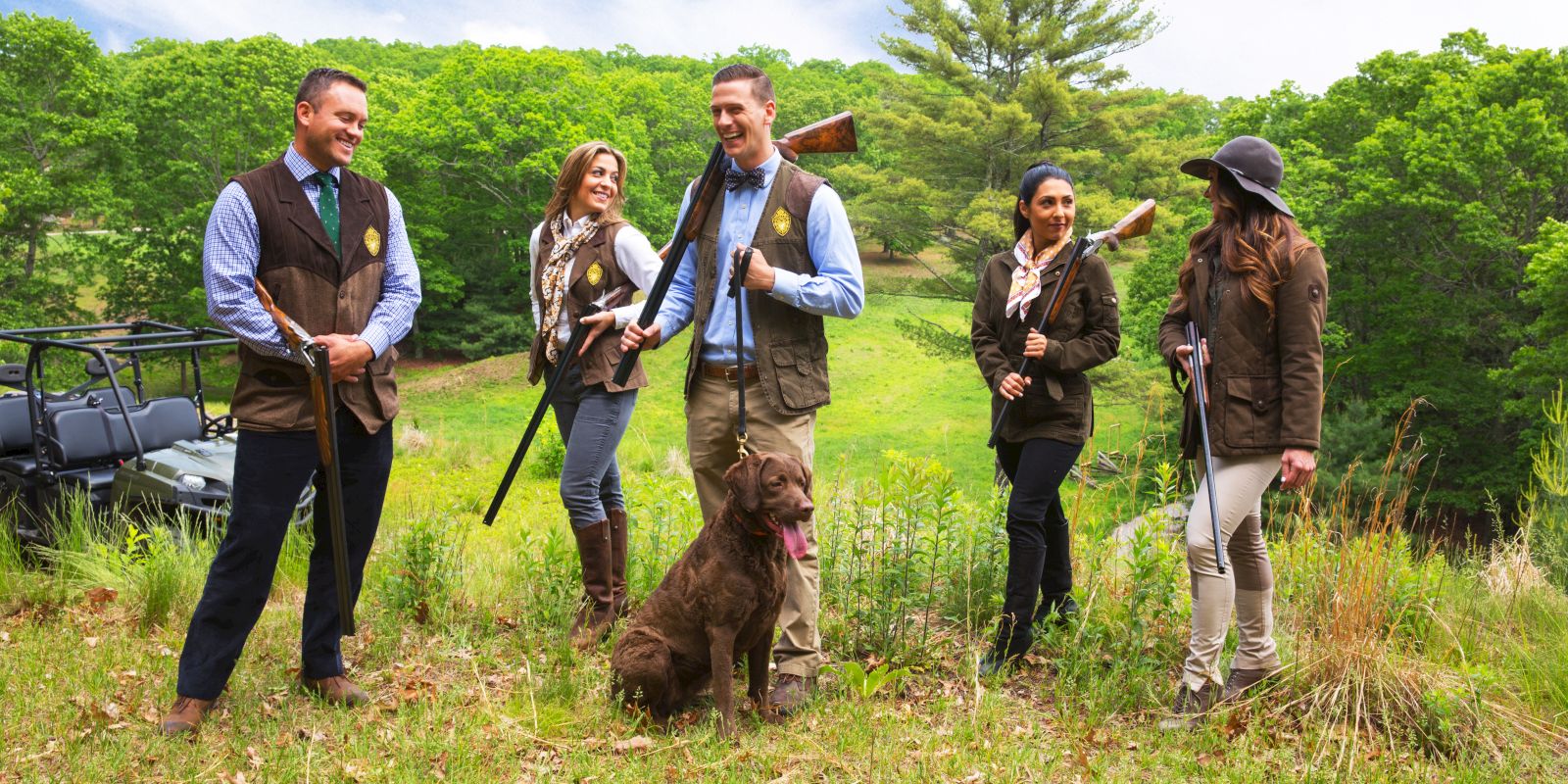 Five people, dressed in outdoor gear, hold rifles next to a dog in a grassy field with a wooded backdrop and vehicle.