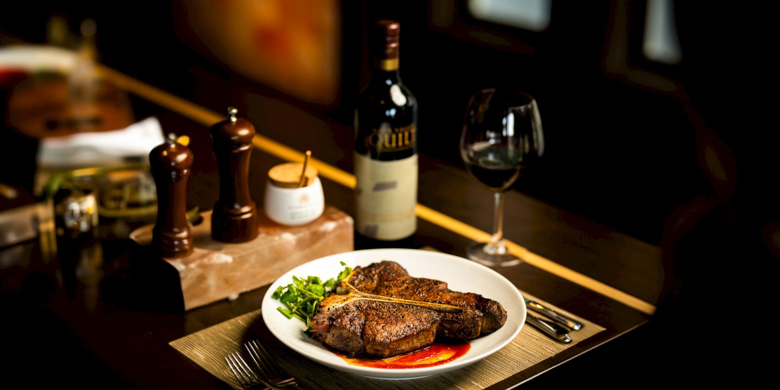 The image shows a plated meal with steak, vegetables, a bottle of red wine, and a glass, on a dark table set for dining.