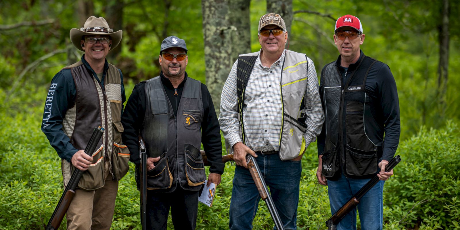 Four men are standing outdoors in front of greenery, holding long firearms and wearing shooting gear, including vests and safety glasses.