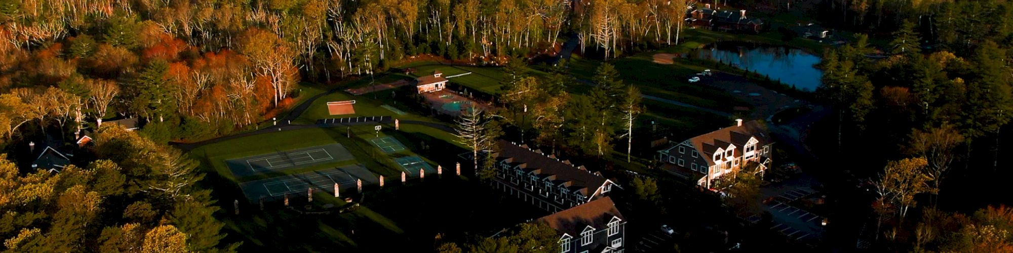 An aerial view of a large estate or resort surrounded by dense forest, featuring multiple buildings, tennis courts, and a small lake in the distance.