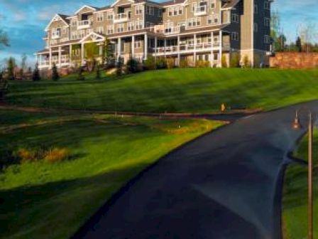 A large, grey multi-story building sits atop a hill, surrounded by green grass and a winding driveway, under a blue sky scattered with clouds.