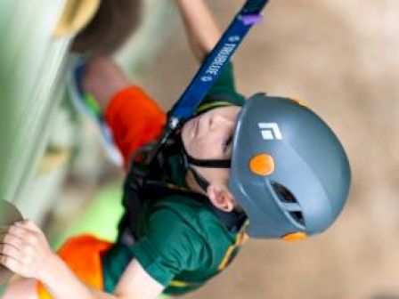 A child wearing a helmet and safety harness climbs a rock wall, focused and reaching for the next handhold.